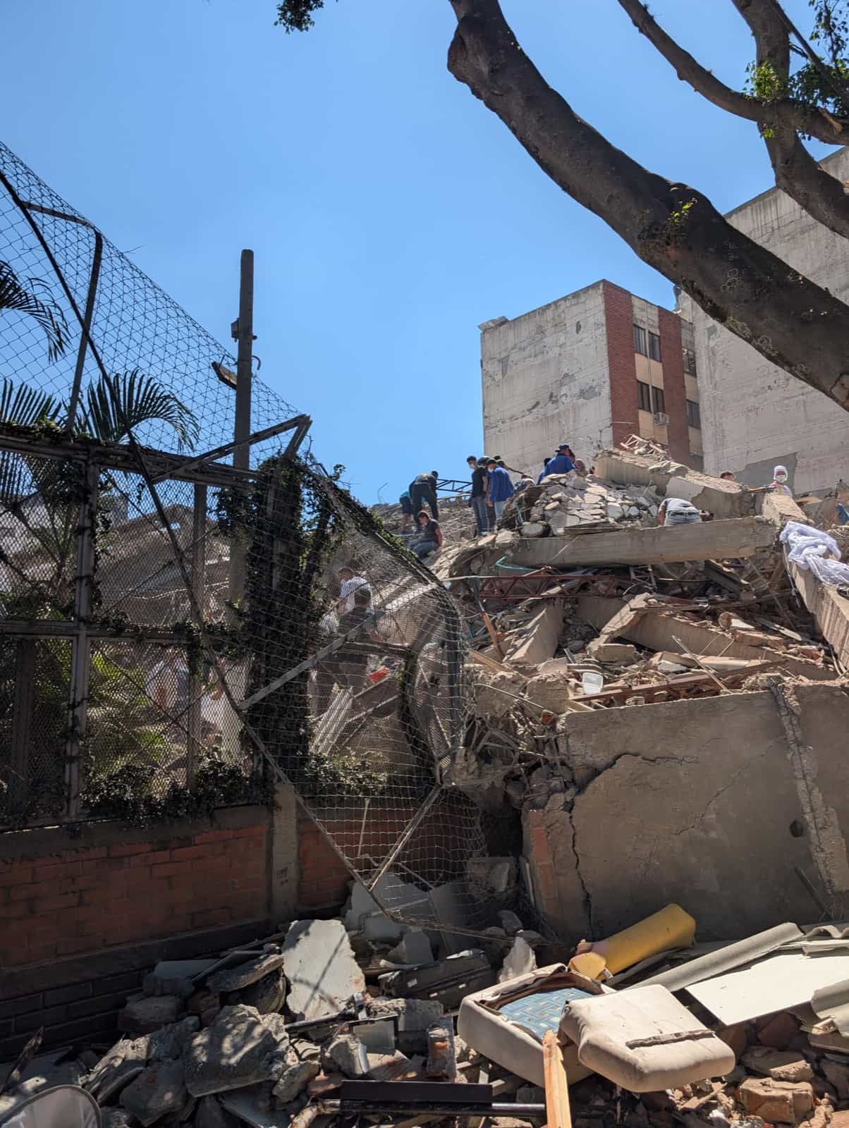 People searching the rubble of a collapsed building. A mattress and broken furniture lie among the concrete and twisted reinforcing bar.