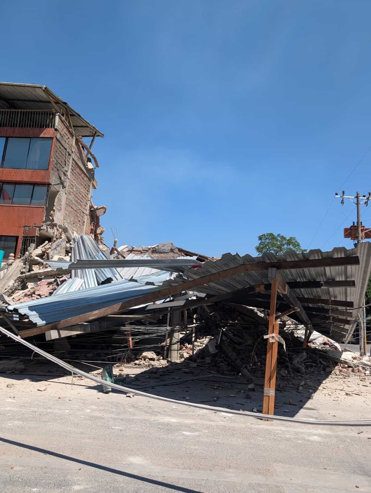 A brick building with one corner collapsed, its corrugated metal roof buckled down onto the street below.