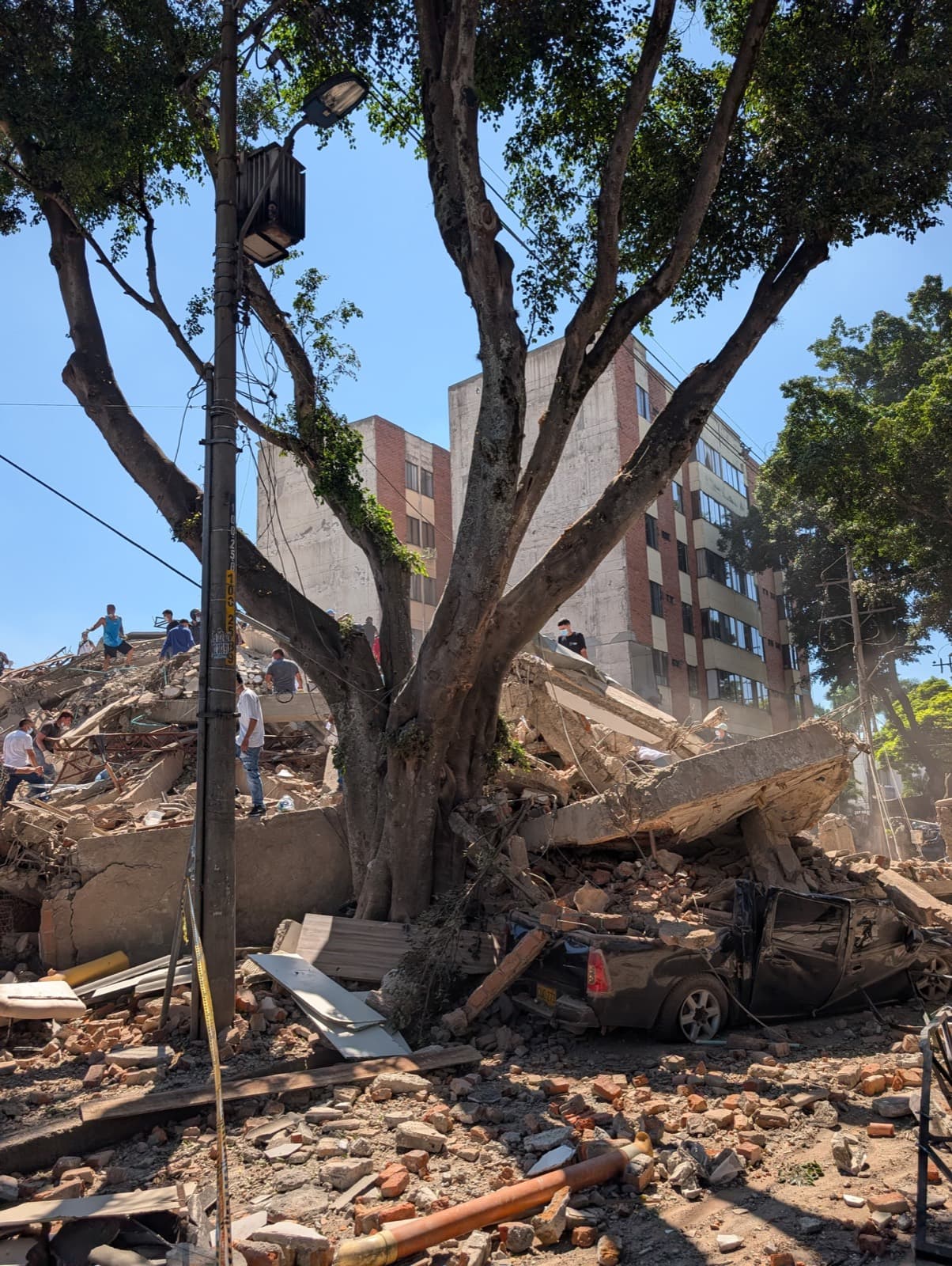 The rubble of a collapsed building spilling into the street, with people standing on top of it and a car crushed beneath a fallen concrete slab. An undamaged apartment block stands behind.