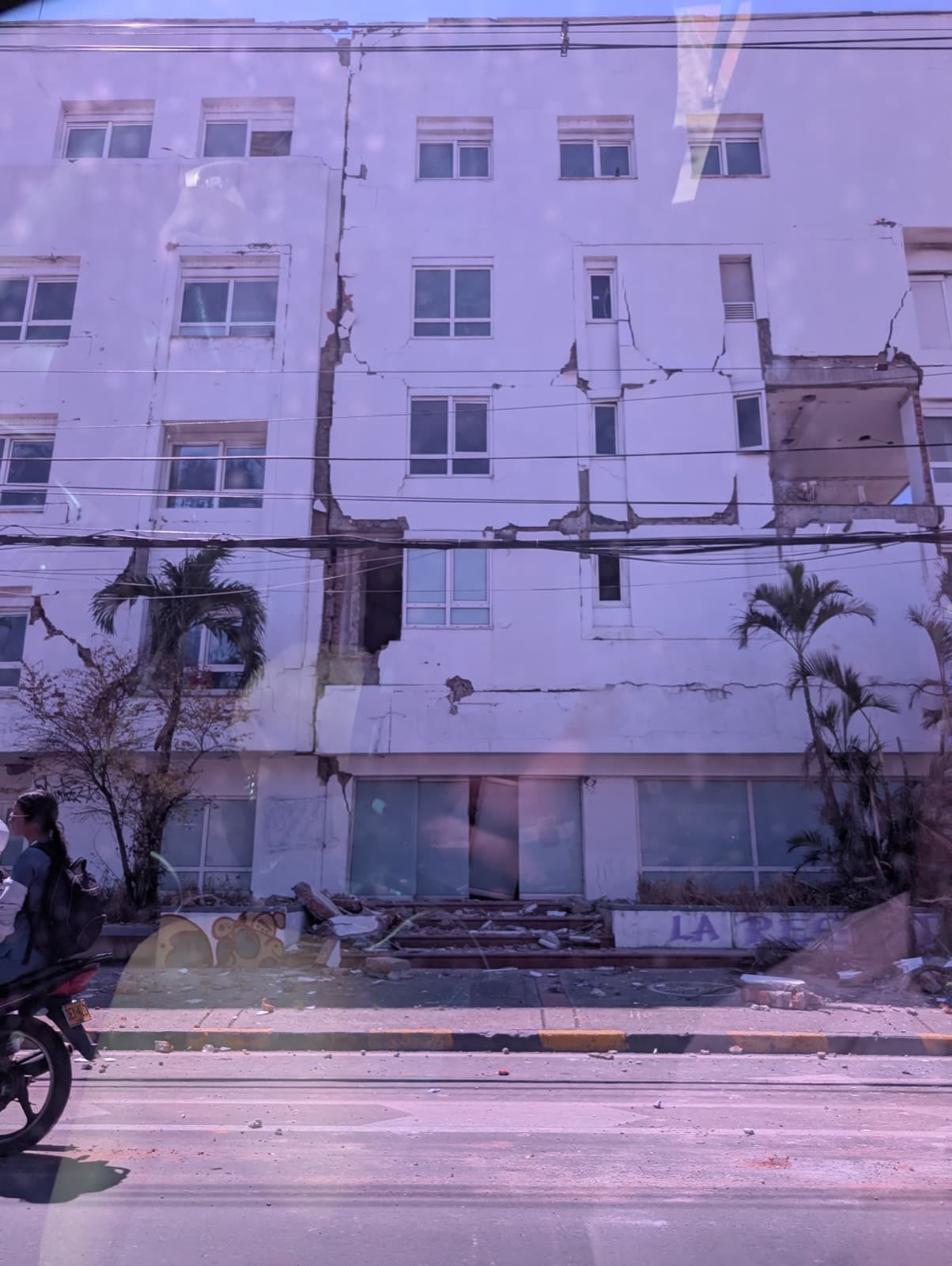 The facade of a white apartment building split by deep structural cracks running floor to floor, photographed from a passing vehicle.