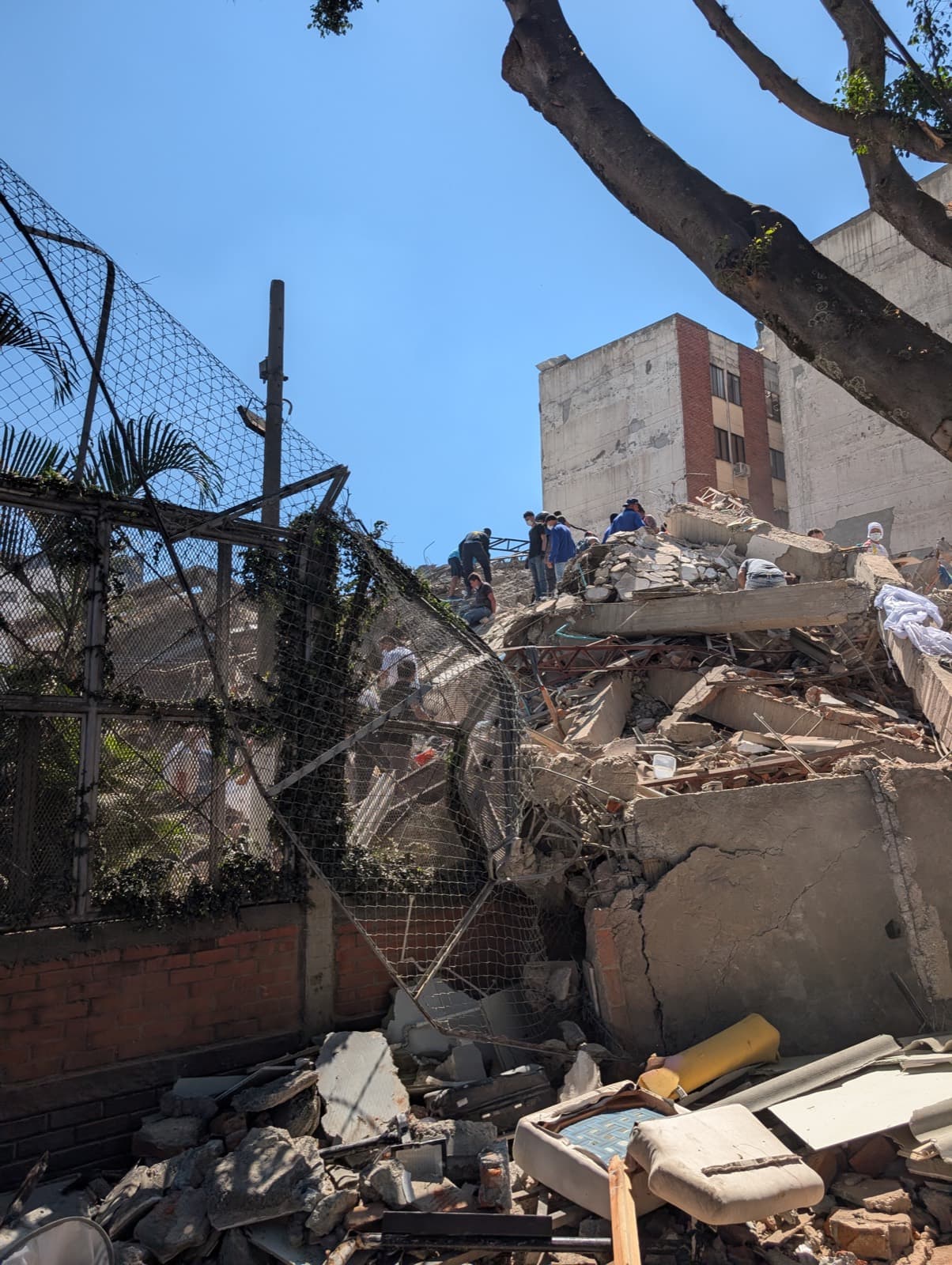 People searching the rubble of a collapsed building. A mattress and broken furniture lie among the concrete and twisted reinforcing bar.