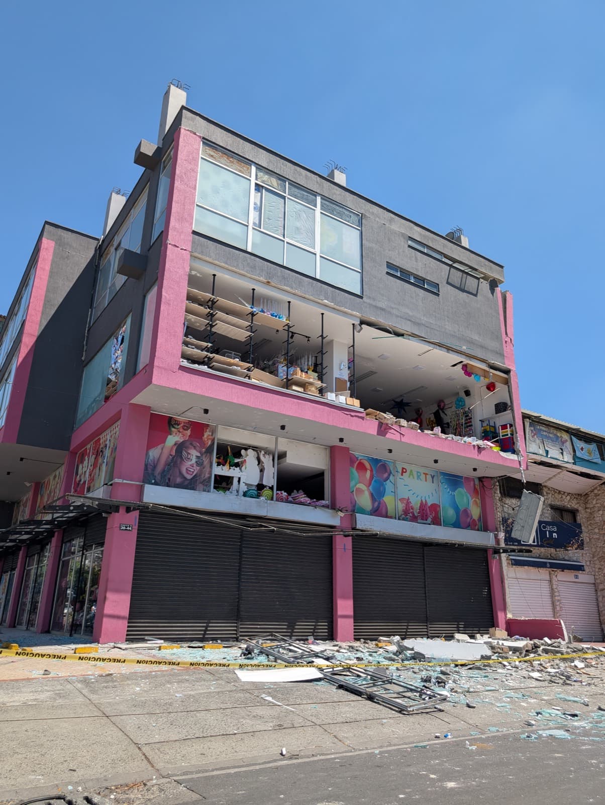 A shop front sheared open by the quake, its upper floors exposed to the street with stock still on the shelves. Broken glass and caution tape cover the pavement.